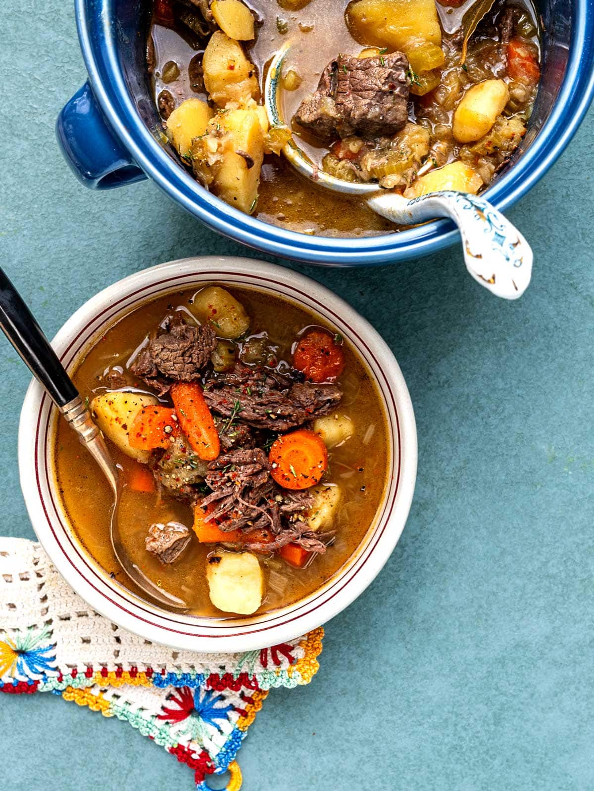 A bowl of soup with shredded beef, carrots and potatoes next to a larger pot of soup.