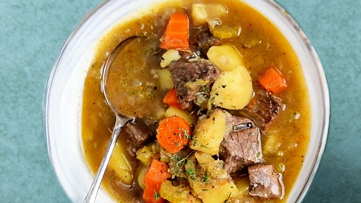 A bowl of beef and vegetable soup with tender beef, potatoes and carrots and a spoon.