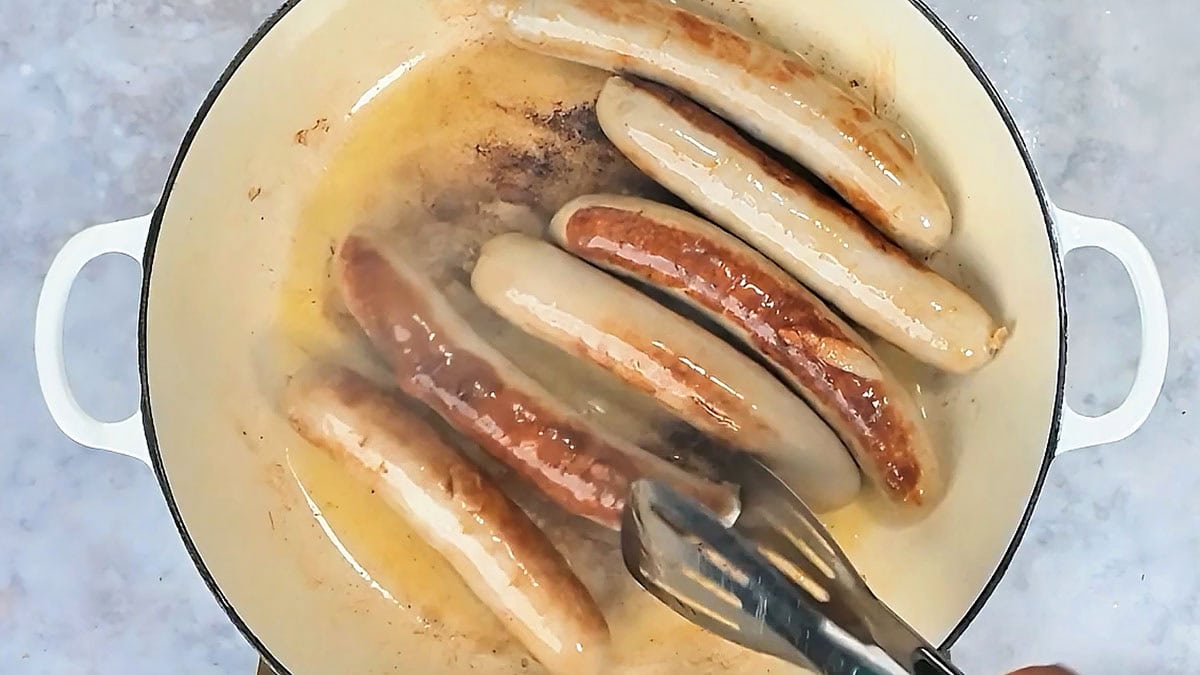 Bratwurst being turned with tongs as they brown in oil in a skillet