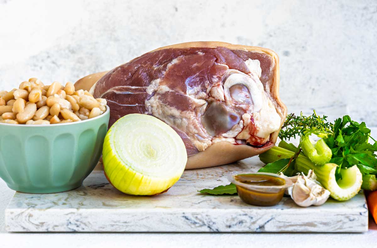 Ingredients for ham and bean soup: a ham hock, white beans, onion, celery, garlic, herbs, stock and oil arranged on a marble board.
