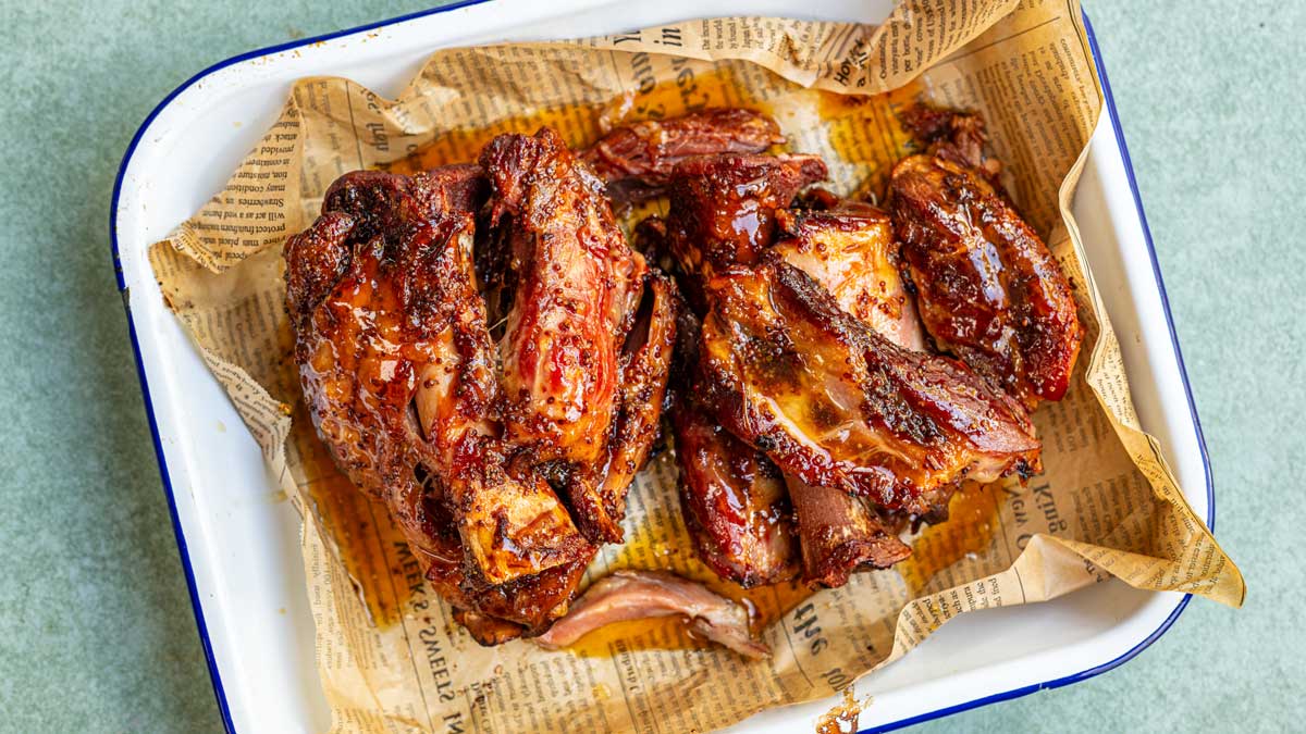 Glazed braised ham hocks, dark and sticky with maple mustard glaze, in a white enamel tray lined with paper.