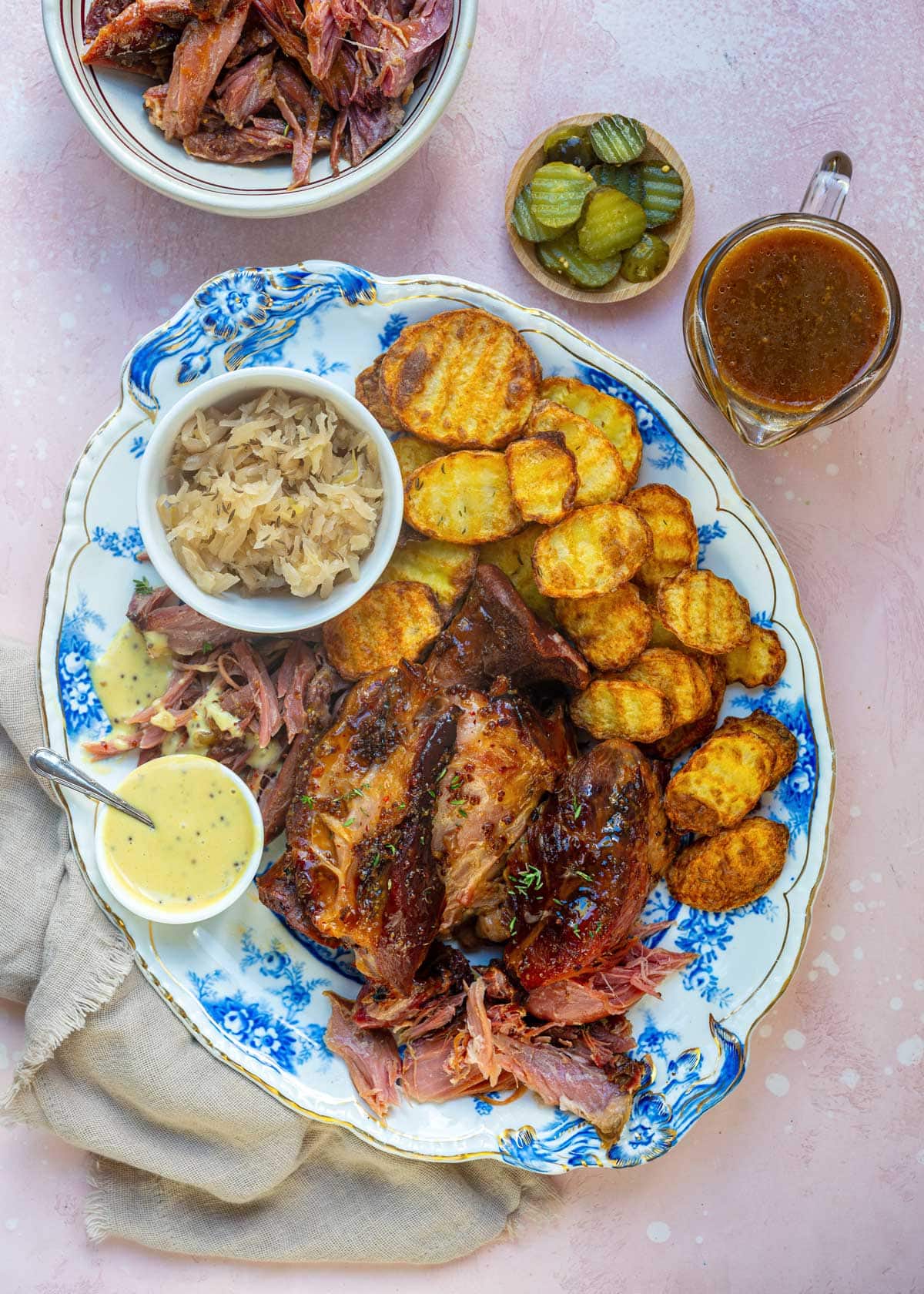 Braised ham hock served on a blue and white platter with crispy potatoes, sauerkraut, mustard sauce, pickles and a jug of gravy.