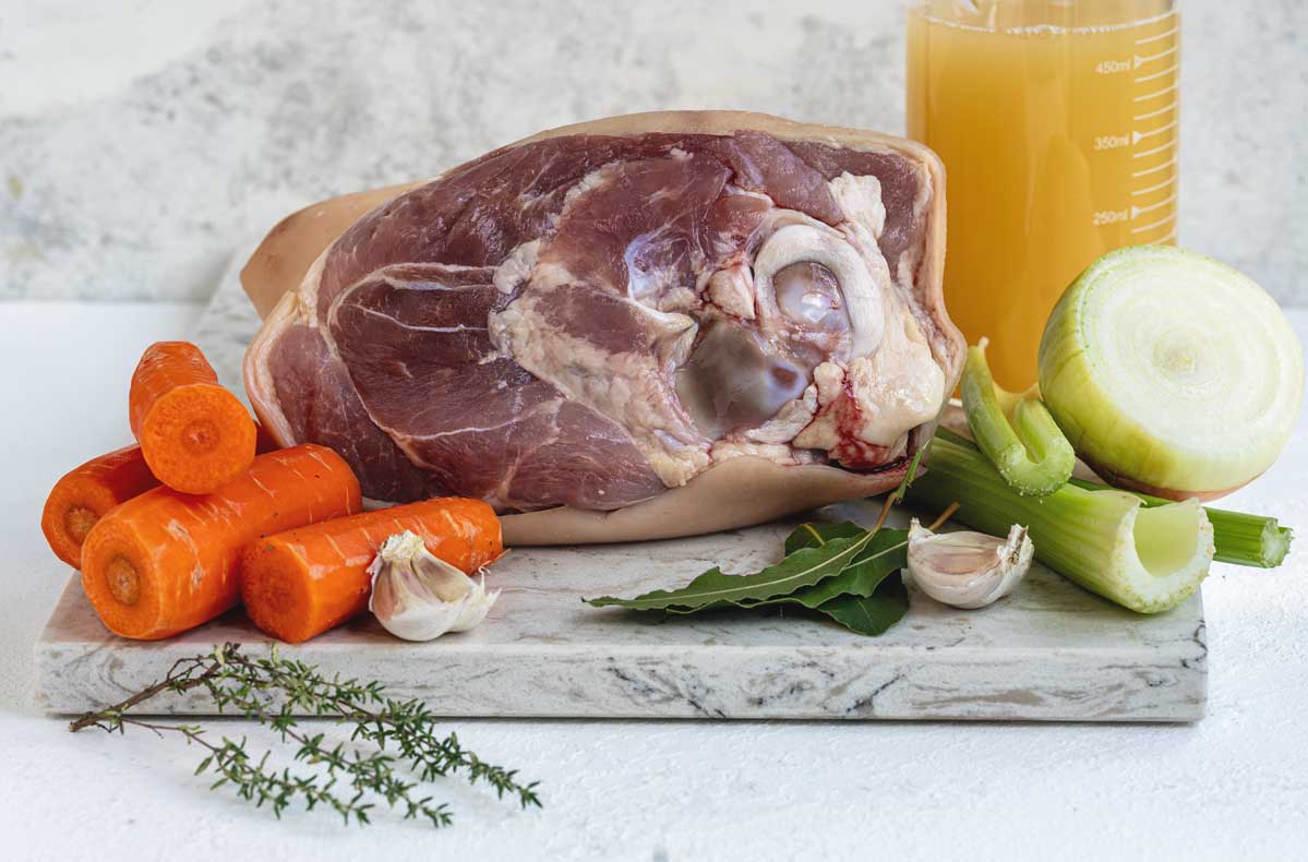 Ingredients for braised ham hock: a raw ham hock on a marble board with carrots, celery, onion, garlic, bay leaves and thyme, and a jar of stock behind.