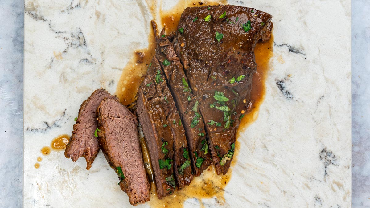 Slices of braised brisket topped with fresh herbs and pan juices on a marble surface.