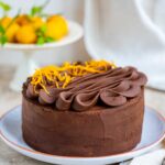 A frosted chocolate orange cake topped with piped frosting swirls and zest, sitting on a white plate with a bowl of oranges in the background.