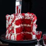 A red velvet cake with a slice removed, decorated with white meringue bones and edible fake blood, with a kitchen knife plunged into the top.