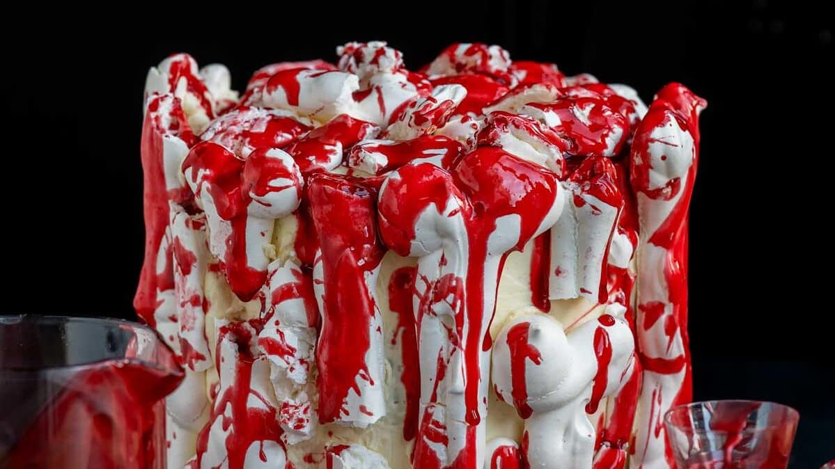 A white cake decorated with meringue bones and drizzled with bright edible fake blood, sitting on a plate next to a pitcher and a glass of red liquid.