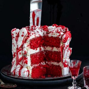 A red velvet cake with a slice removed, decorated with white meringue bones and edible fake blood, with a kitchen knife plunged into the top.