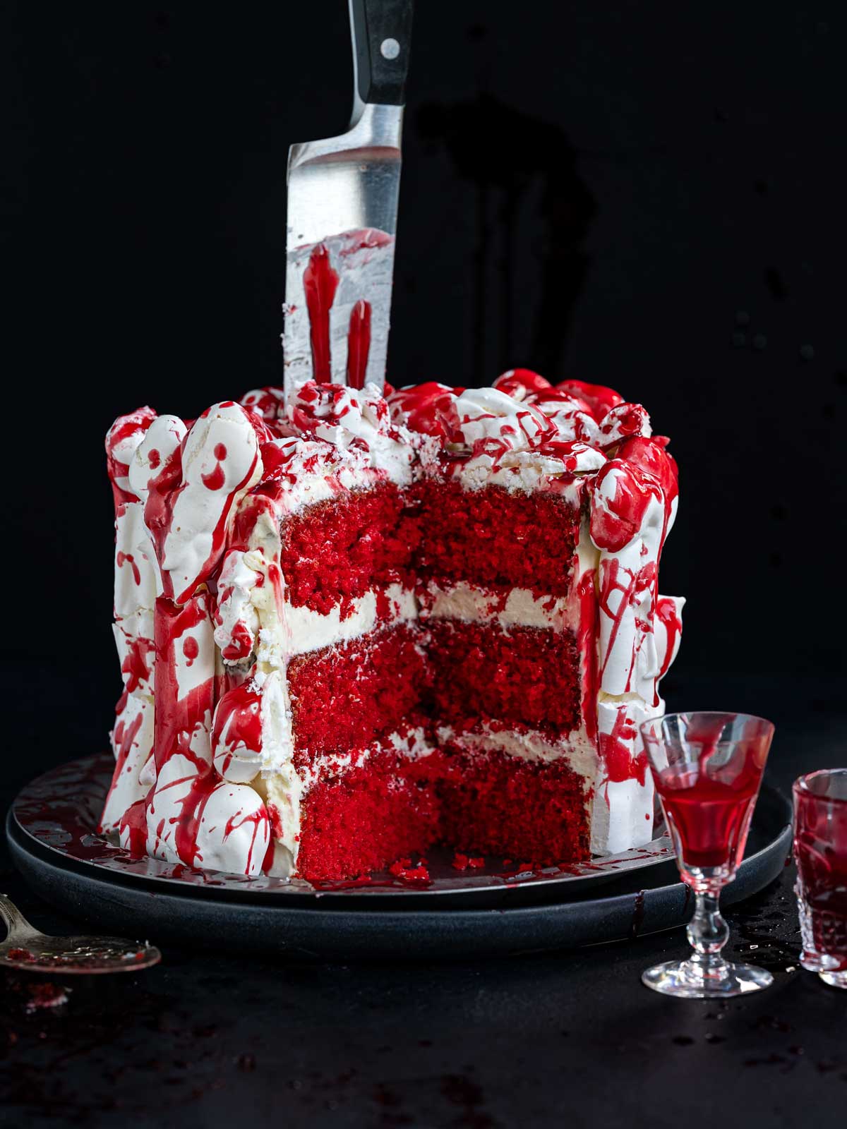 A red velvet cake with a slice removed, decorated with white meringue bones and edible fake blood, with a kitchen knife plunged into the top.