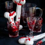 Halloween meringue bones splattered with edible fake blood, served in skull-shaped glasses alongside a measuring pitcher of dark red liquid.