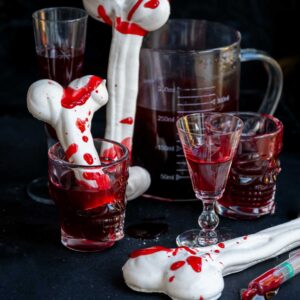 Halloween meringue bones splattered with edible fake blood, served in skull-shaped glasses alongside a measuring pitcher of dark red liquid.