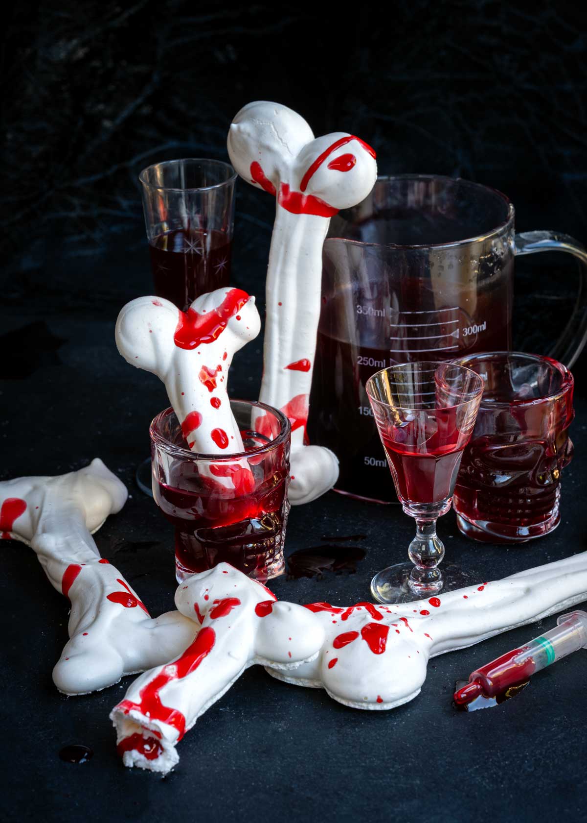 Meringue bones splattered with edible fake blood, served alongside glasses filled with dark red liquid and a plastic syringe.