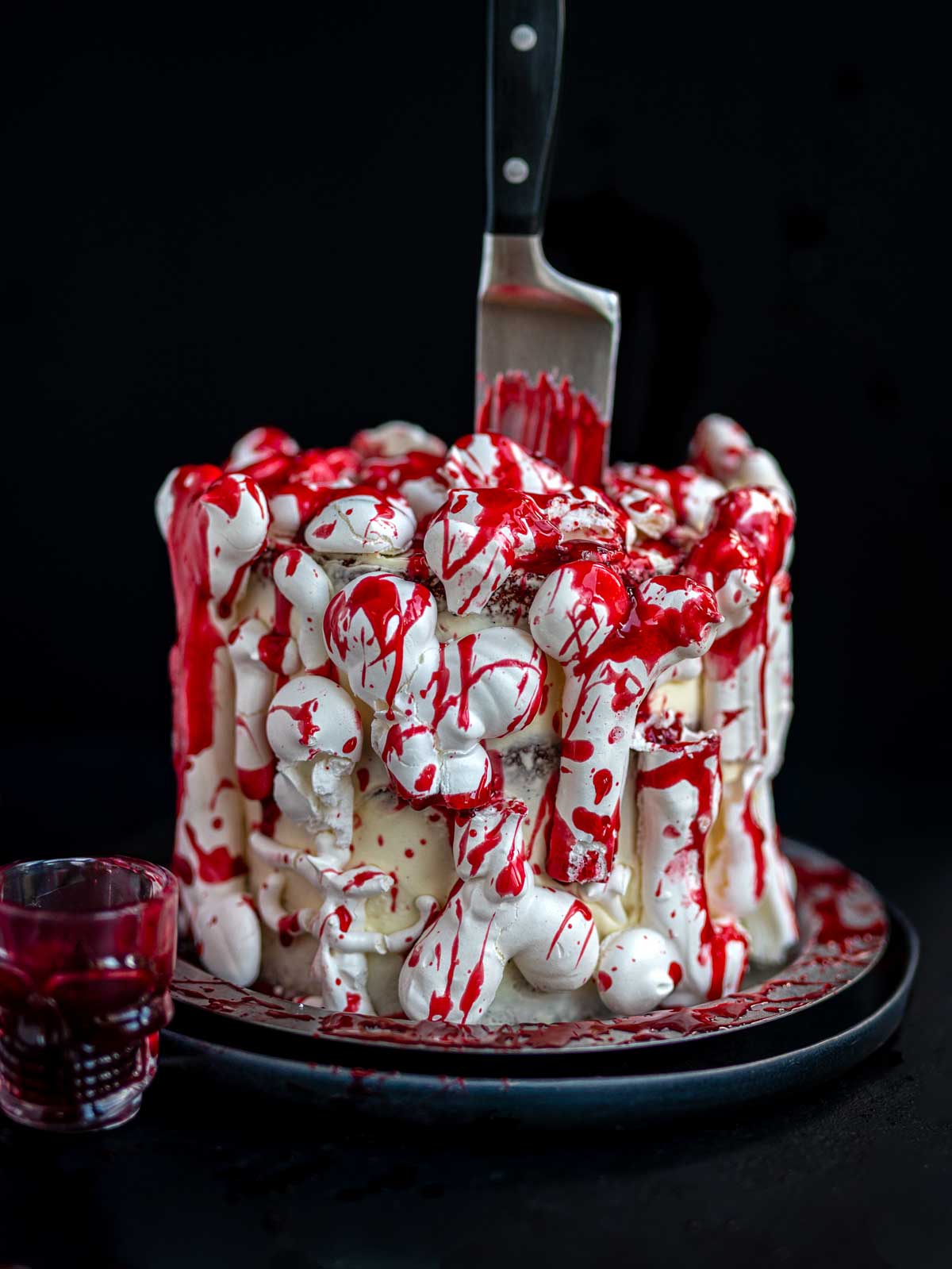 A cake covered in white meringue bones and edible fake blood drips, with a large knife stuck into the top and a small skull-shaped glass of red liquid beside it.