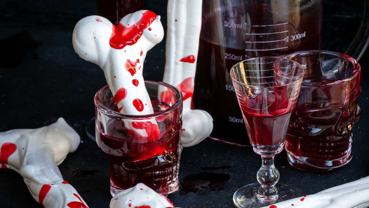 A white meringue bone splattered with edible fake blood sits in a skull-shaped shot glass filled with dark red liquid, next to a laboratory beaker and a crystal glass.