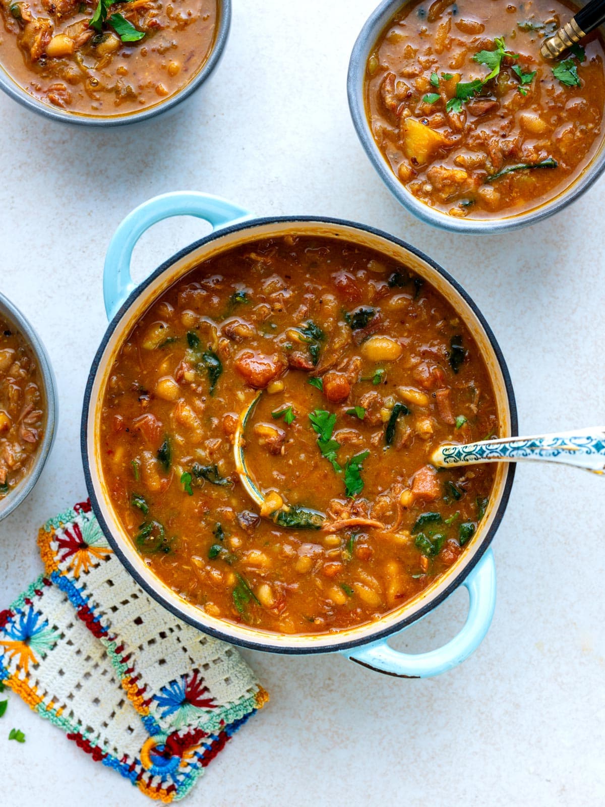 A large blue pot filled with ham and bean soup, surrounded by two smaller bowls of the same dish and a crocheted coaster.