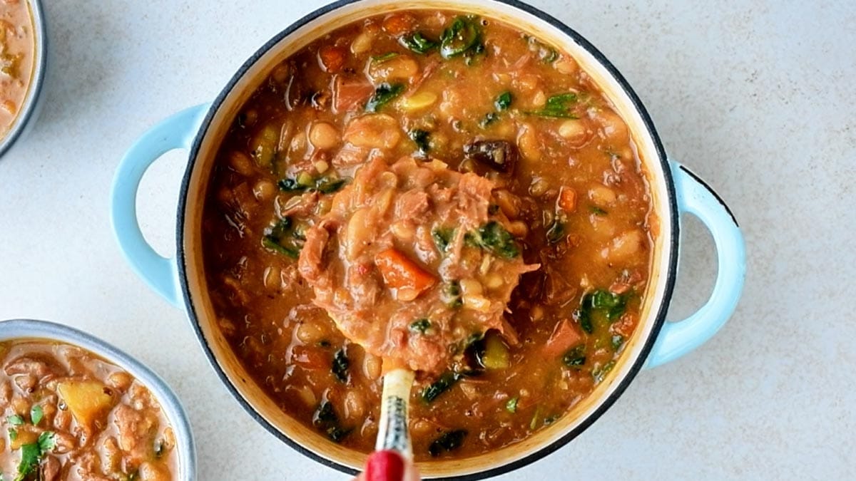 A ladle lifts a scoop of hearty ham and bean soup from a blue enameled pot.