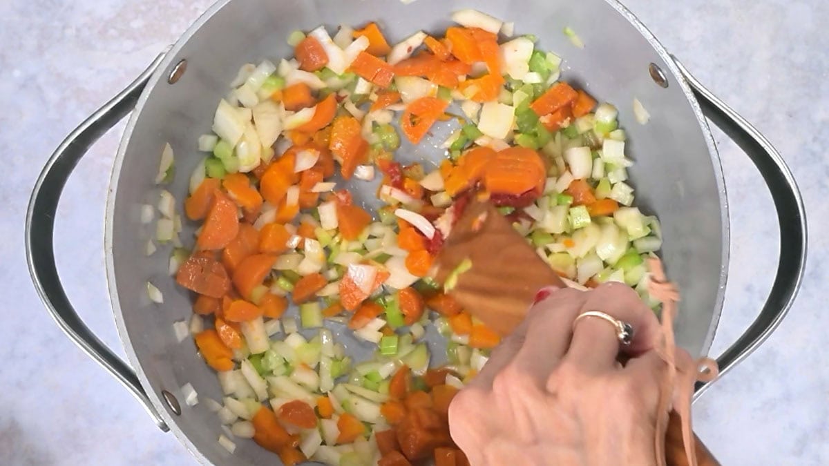 A person uses a wooden spatula to stir chopped carrots, onions, and celery for ham and bean soup in a large pan.