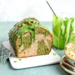 A loaf of marble matcha banana bread topped with green glaze, next to a glass of green glaze and a plate with a slice of cake.
