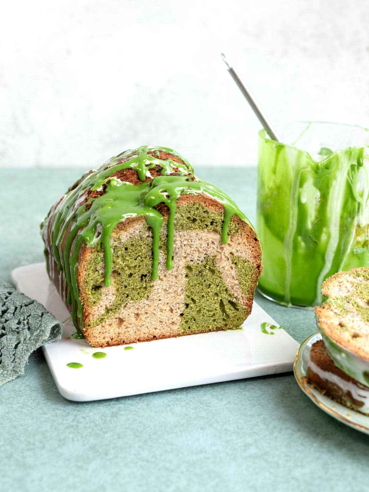 A loaf of marble matcha banana bread topped with green glaze, next to a glass of green glaze and a plate with a slice of cake.