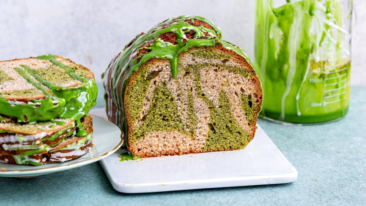 A loaf of sliced green and beige marbled matcha banana bread topped with green glaze, with additional slices on a small plate to the left.