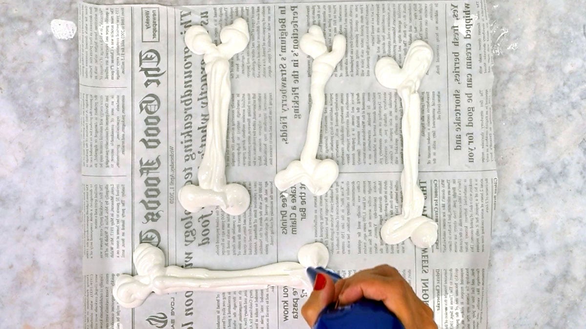 A hand piping white meringue bones with a blue piping bag onto a parchment-lined baking sheet.