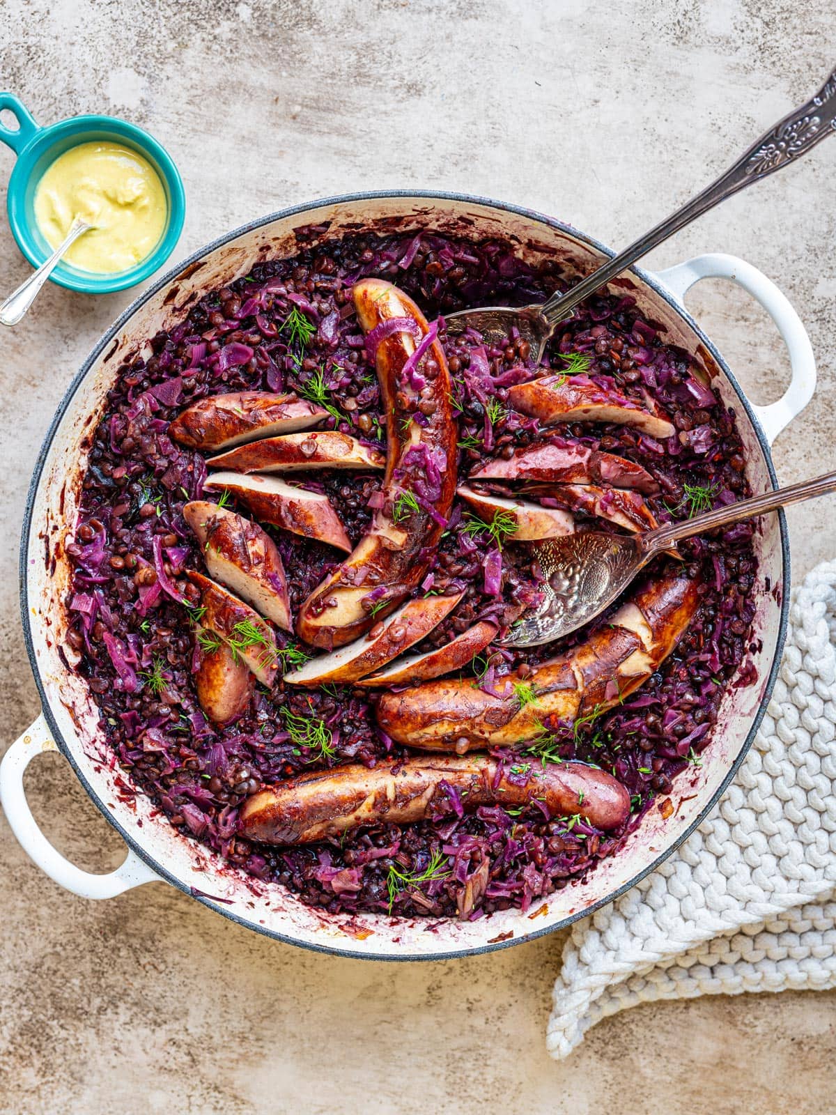 Bratwurst casserole in a cream braiser – browned bratwurst, some sliced, on a bed of lentils and braised red cabbage, with serving spoons and a small pot of mustard