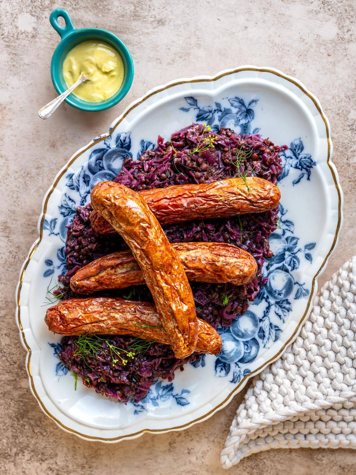 Grilled bratwurst served over braised red cabbage and lentils on a blue and white floral platter, with a small pot of mustard