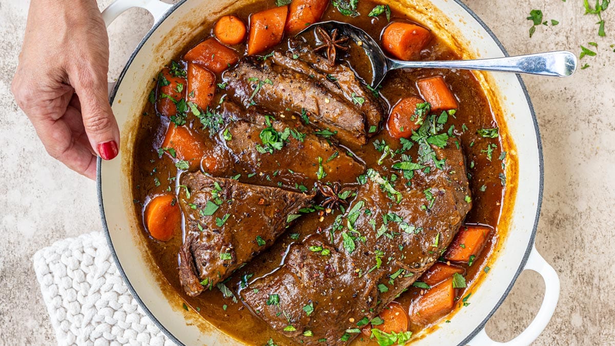 A hand holding the handle of a white Dutch oven filled with sliced braised brisket and carrots in a dark gravy, garnished with fresh herbs and star anise.