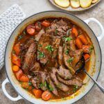 Sliced braised brisket and carrots in a rich brown gravy in a white Dutch oven, garnished with fresh herbs and star anise, with a plate of roast potatoes on the side.