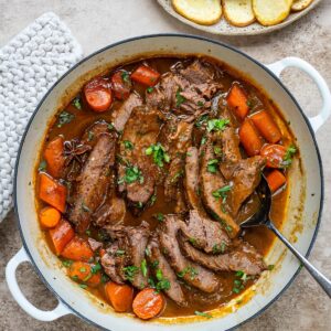 Sliced braised brisket and carrots in a rich brown gravy in a white Dutch oven, garnished with fresh herbs and star anise, with a plate of roast potatoes on the side.