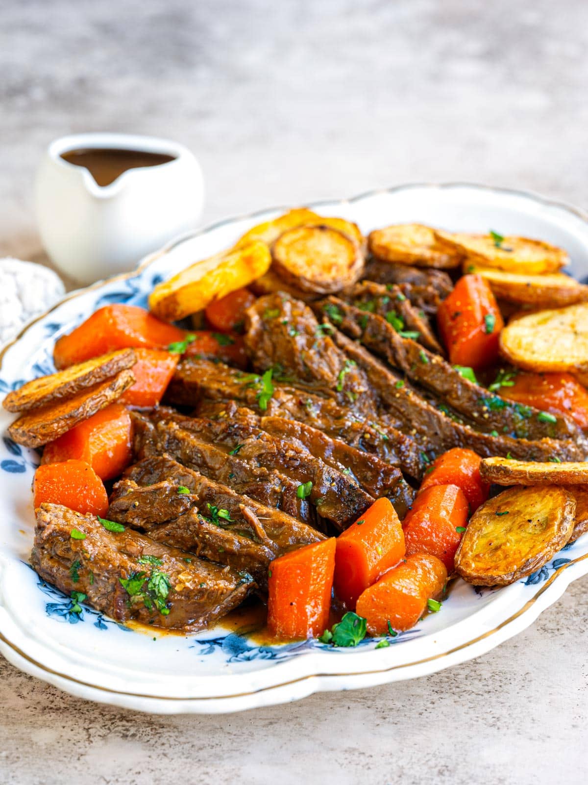 Sliced braised brisket on a blue and white platter with carrots and roast potatoes, with a small jug of gravy on the side.