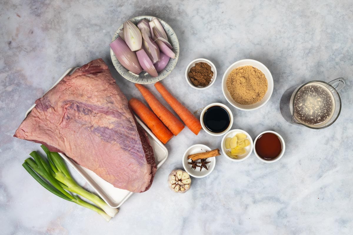 Ingredients for oven braised brisket on a marble surface: a raw beef brisket, shallots, carrots, green onions, a head of garlic, ginger, star anise, a cinnamon stick, brown sugar, soy sauce, fish sauce, five spice and a jug of beef broth.