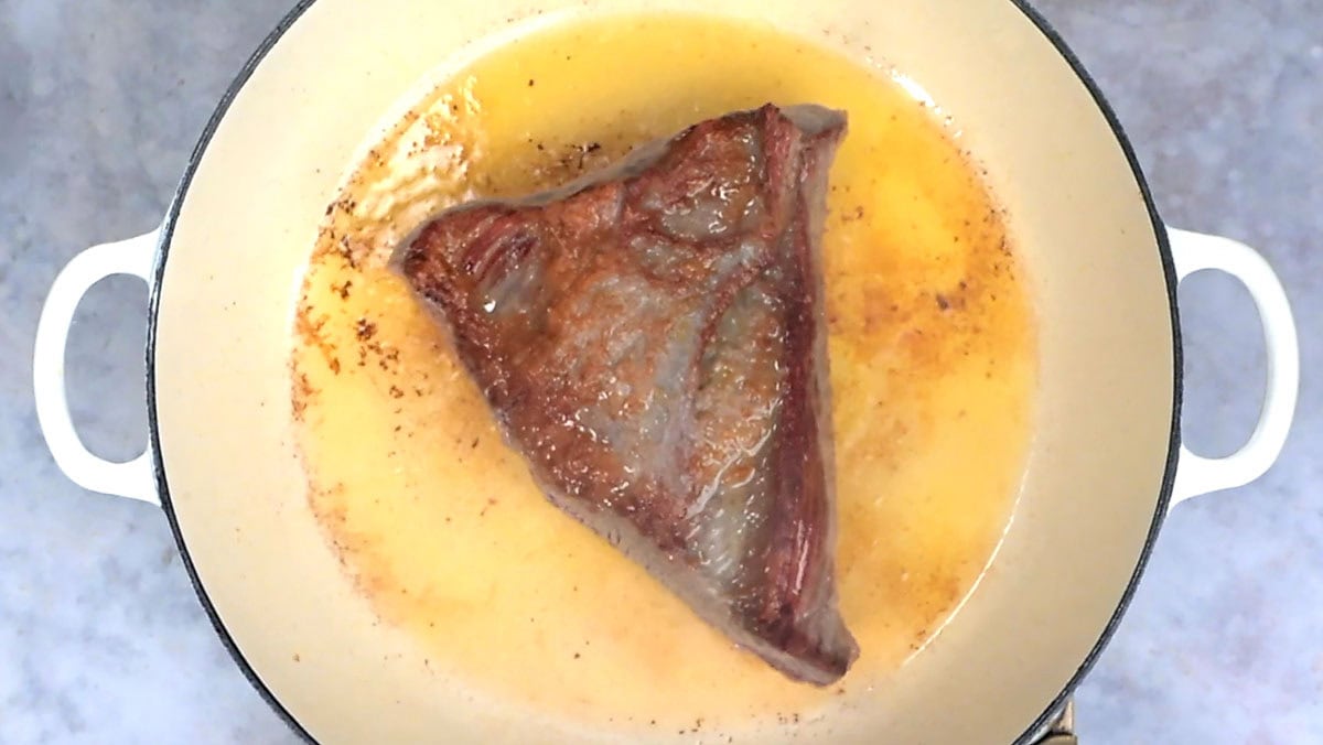 Half a raw beef brisket searing in hot oil in a white Dutch oven.