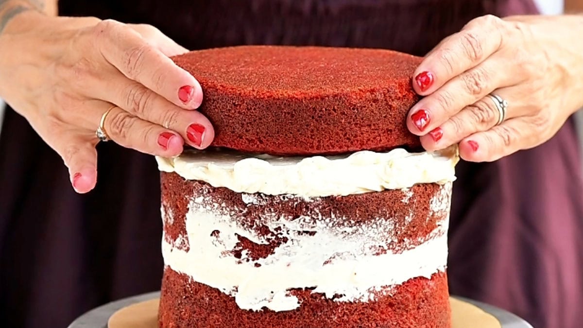 Hands place a red velvet cake layer on top of a frosted stack decorated with meringue bones.