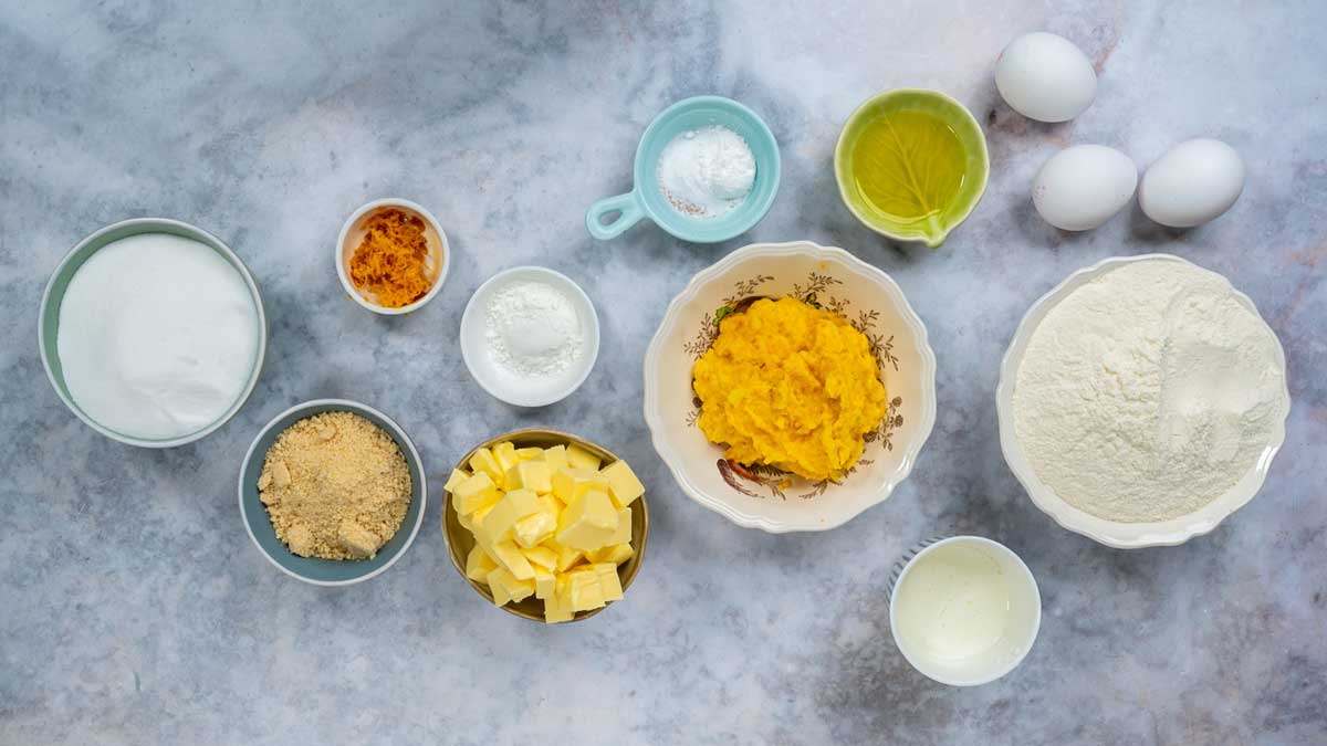 Baking ingredients for a chocolate orange cake arranged on a grey surface, including bowls of flour, sugar, cubed butter, mashed pumpkin, eggs, and citrus zest.