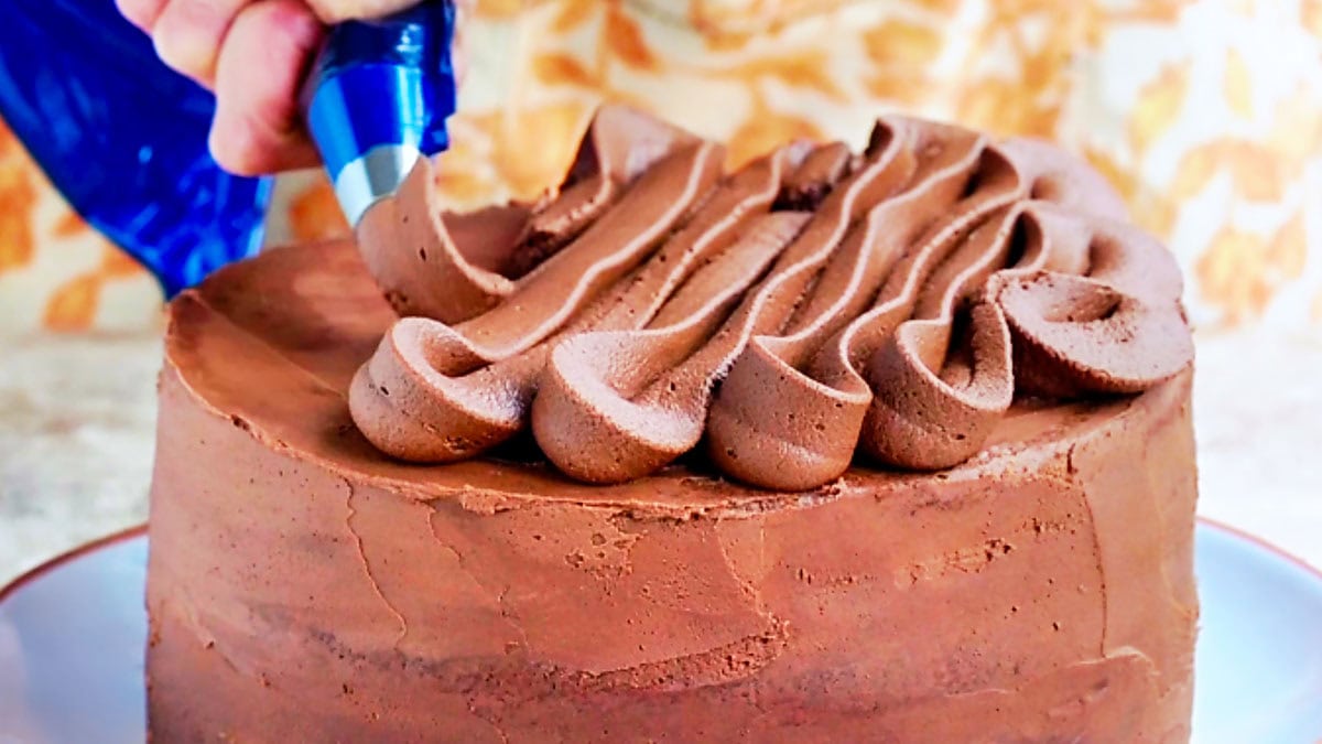 A hand uses a piping bag to add decorative swirls of chocolate frosting to the top of a chocolate orange cake.