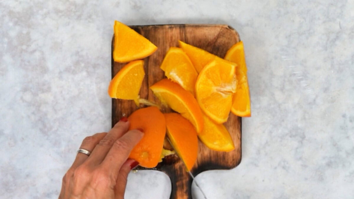 A hand uses a knife to cut orange segments on a wooden cutting board for a chocolate orange cake.