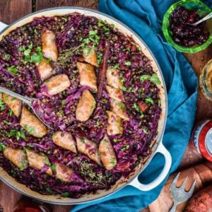 Overhead view of bratwurst casserole with lentils and red cabbage in a shallow braiser, with a glass of German beer, a can of beer and a dish of cranberry sauce