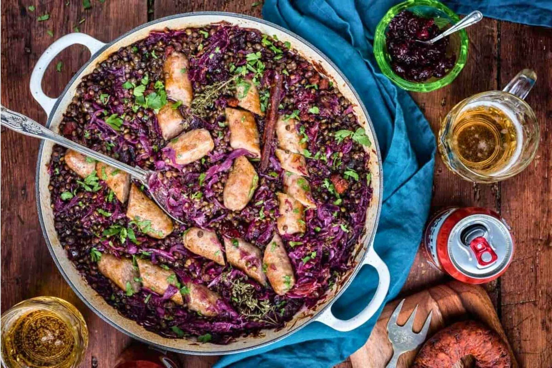 Overhead view of bratwurst casserole with lentils and red cabbage in a shallow braiser, with a glass of German beer, a can of beer and a dish of cranberry sauce