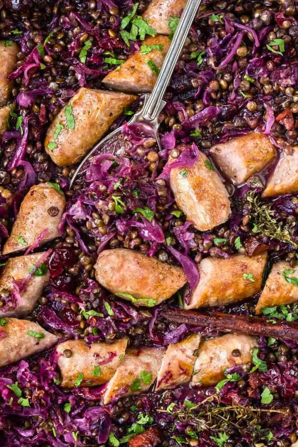 Sliced sausages served with braised red cabbage, lentils, and fresh herbs, with a serving spoon resting in the center.